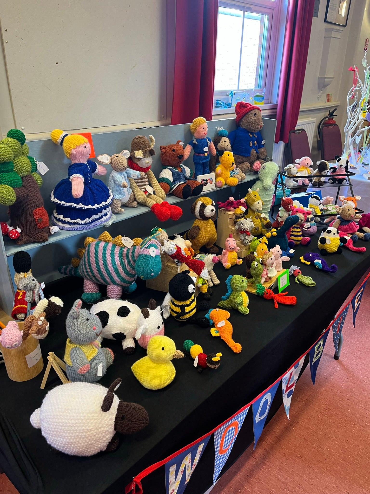 Collection of various hand knitted and crocheted soft toys on a table with handmade bunting showing the makers name
